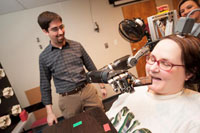 Jan Scheuermann, who has quadriplegia, prepares to take a bite out of a chocolate bar she has guided into her mouth with a thought-controlled robot arm. Research assistants Drs. Brian Wodlinger and Elke Brown watch in the background. (Photo: UPMC)) 