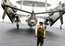 A flight deck crew member stands behind an E-2C Hawkeye aboard the flight deck of the aircraft carrier USS Saratoga during Operation Desert Storm. 